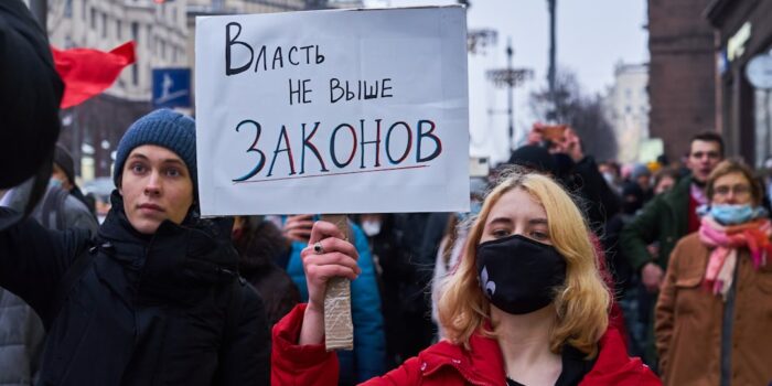 Person mit rotem Mantel hält ein Plakat mit der Aufschrift 'Vlast ne vyshe zakonov' während einer Demonstration.