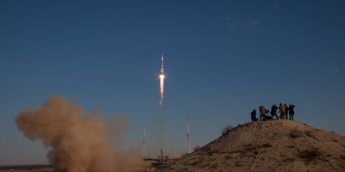 Eine Rakete startet vom Boden, während Zuschauer auf einem Hügel stehen und zuschauen.